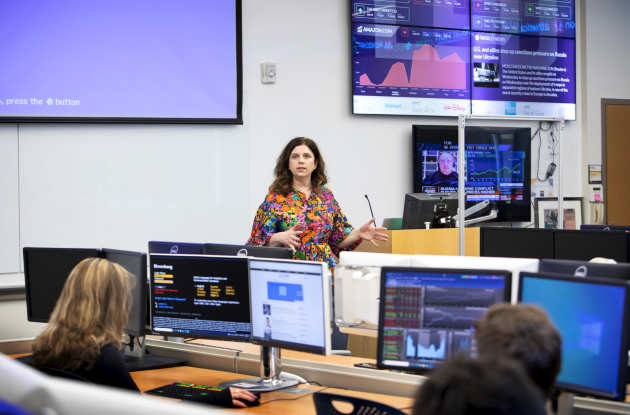 Instructor talking at the front of SMART Lab with computers displaying financal data in the background. 